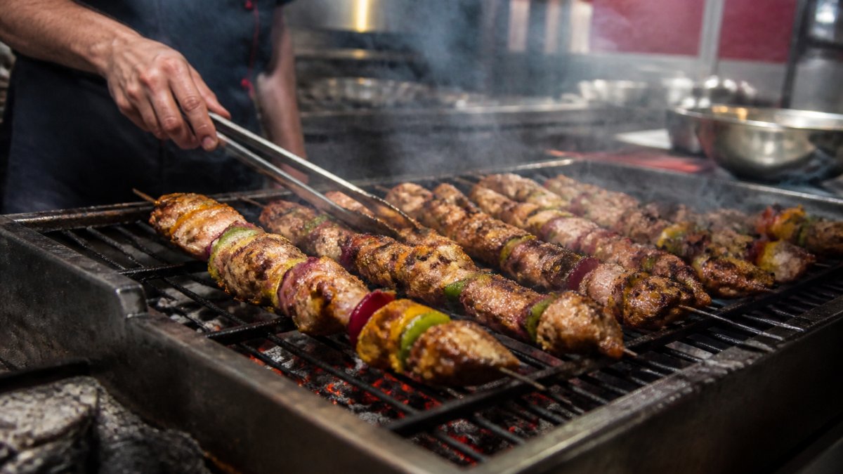 Close-up of skewers of meat and vegetables sizzling over glowing charcoal on a grill, with light smoke rising and a chef's hand turning them with tongs in a busy open kitchen