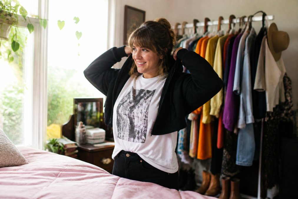 A smiling woman chooses cathartic clothing from a colorful rack in a sunlit bedroom to embrace dopamine dressing.