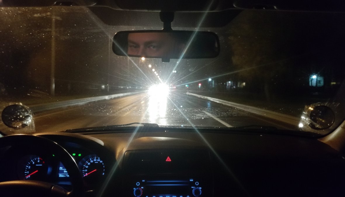 Close-up of a driver's perspective at night, viewed from inside the car cabin, with multiple oncoming headlights creating bright halos and streaks across a wet road surface, dashboard illuminated softly in the foreground