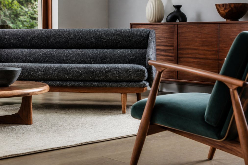 Styled lounge space showcasing a sculptural mid-century modern sofa with tapered wooden legs and textured charcoal upholstery, paired with a retro accent chair in jewel-tone velvet, close-up detail of wooden frame joinery and upholstery texture under warm ambient lighting