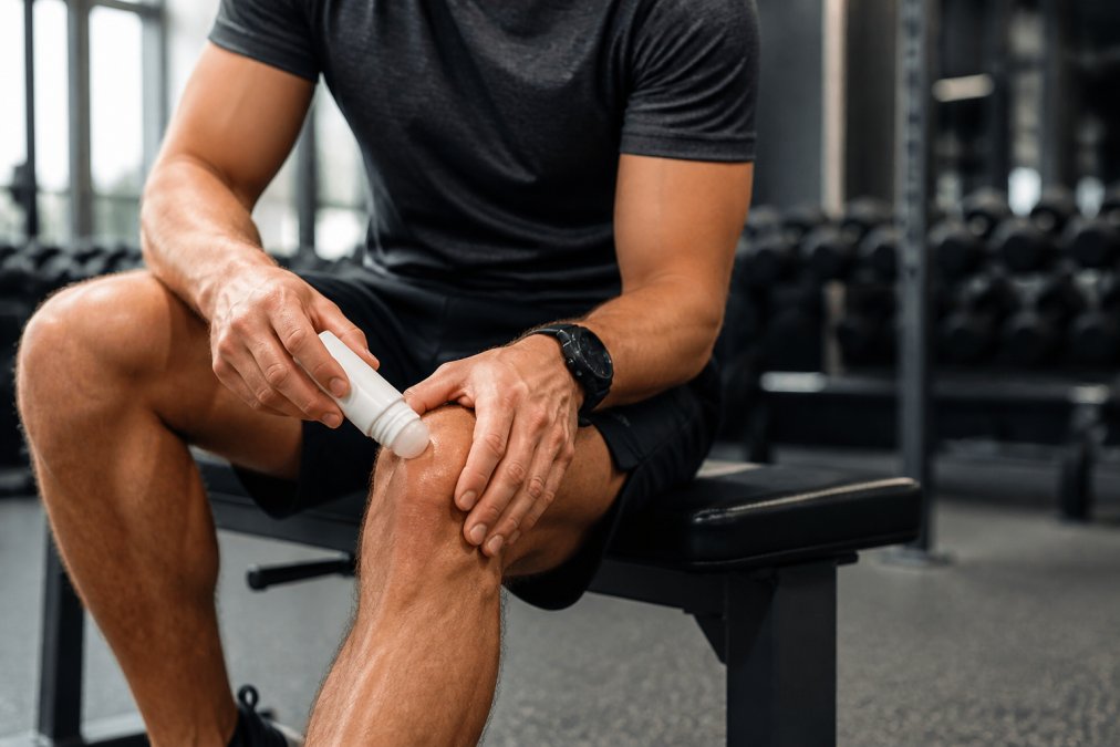 Athlete applying CBD freeze gel roll-on to knee during post-workout recovery session, showing the roll-on applicator in direct contact with skin, athletic wear visible in background with natural gym lighting