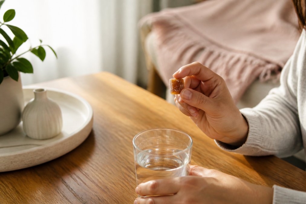 Close-up of hands holding a single CBD gummy over a glass of water on a wooden table in soft morning light, peaceful and calm setting