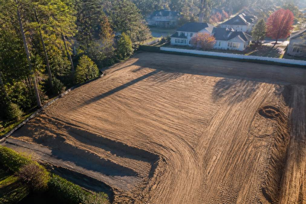 Aerial view of a residential property showing freshly graded earth with compacted soil in geometric patterns, cleared land prepared for construction, morning sunlight casting shadows across the prepared site