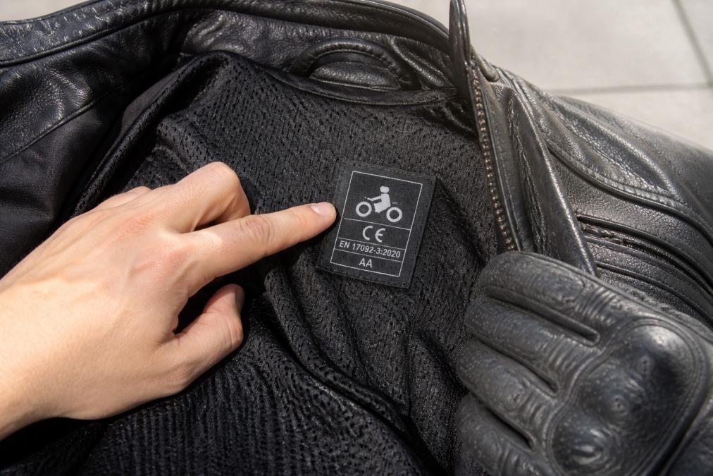Close-up of a rider's gloved hand pointing to a woven CE label and tag sewn inside the lining of a leather motorcycle jacket, showing the EN 17092 markings in natural daylight