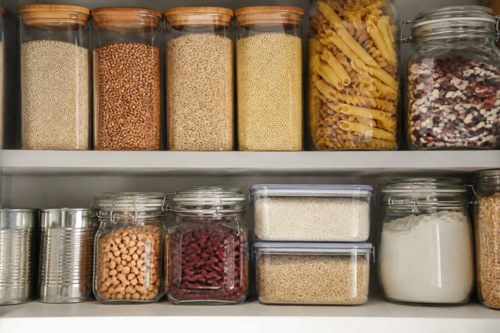 Overhead view of an organized pantry shelf displaying gluten-free staples: containers of quinoa, brown rice, buckwheat, millet, canned beans, certified gluten-free pasta, and whole food ingredients arranged in clear glass containers with labels