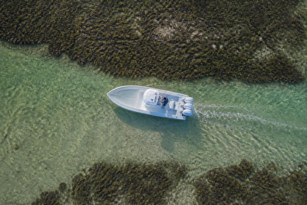 Overhead view of a shallow water fishing boat navigating through narrow flats with visible sand bottom, showing the slim profile and low draft design cutting through shallow water