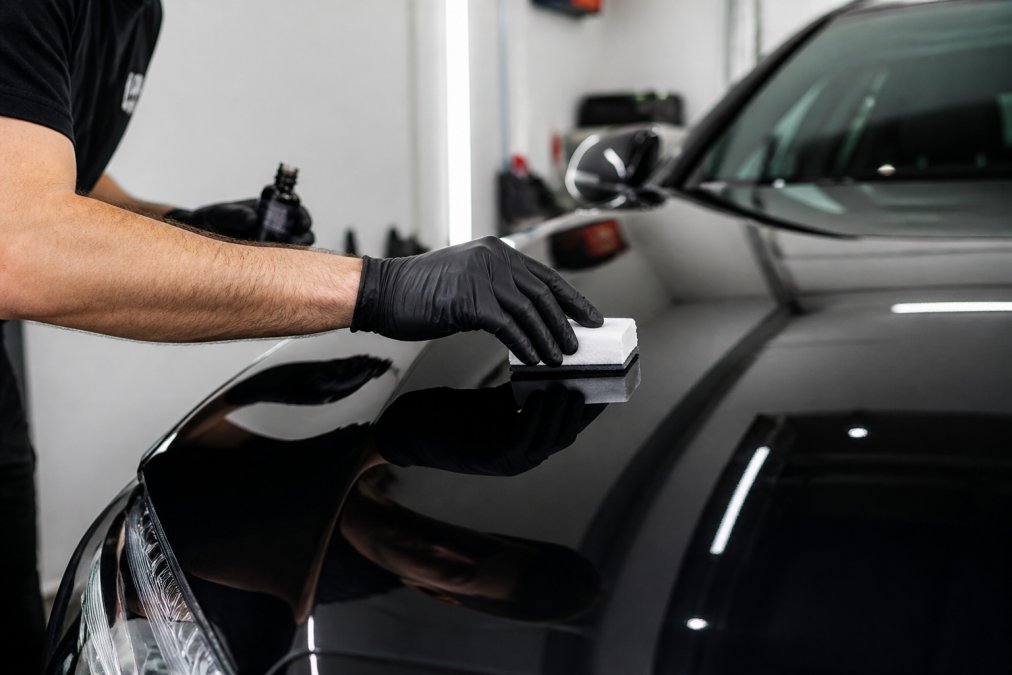 Close-up of a technician's gloved hands applying a ceramic coating to the glossy black hood of a luxury sedan inside a clean, well-lit detailing studio