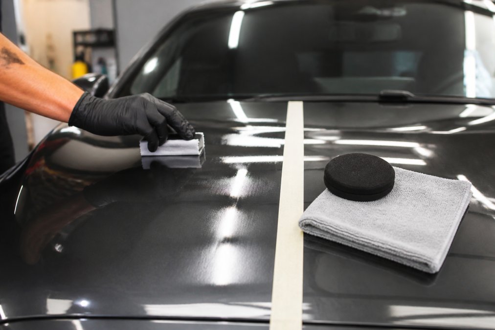 Close-up of a detailer's gloved hand applying ceramic coating to a glossy dark car hood with a microfiber applicator in a bright detailing bay