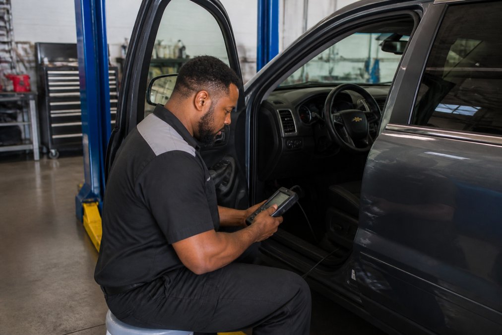 An ASE-certified automotive technician in a branded uniform using a handheld diagnostic scanner plugged into the OBD-II port of a Chevrolet SUV inside a clean, well-lit auto repair bay, with tool cabinets and a lift visible in the background