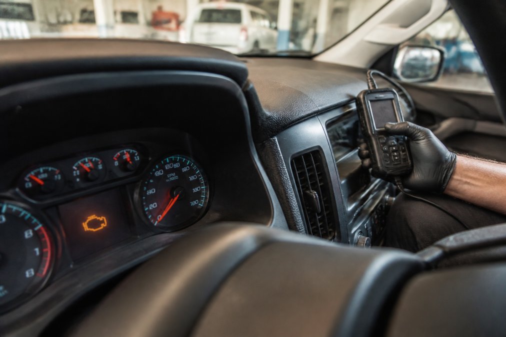 Close-up of a check engine light illuminated on the dashboard instrument cluster of a Chevrolet vehicle, with a mechanic's hand visible in the background holding a diagnostic scanner tool, shot in warm workshop lighting