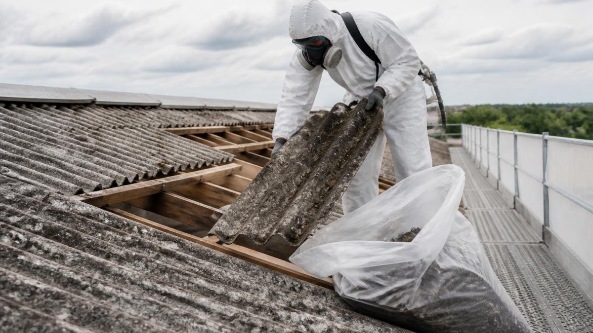 Certifikovaný pracovník v ochranném oděvu a respirátoru provádějící bezpečnou demontáž azbestového materiálu ze střechy, se speciálními pytly a hermetickým vybavením