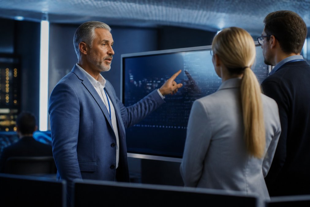 A security architect pointing at a large monitor displaying a cryptographic key authorization workflow in a modern enterprise operations center with blue ambient lighting