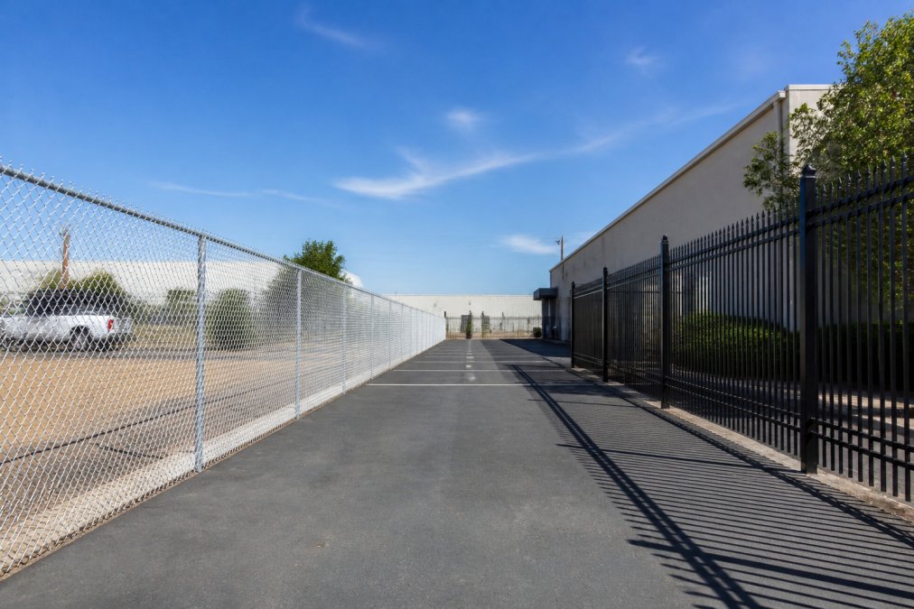A wide shot of a commercial parking lot where a galvanized chain link fence runs along one side and a black ornamental iron fence borders the other, with a warehouse and clear blue sky in the background
