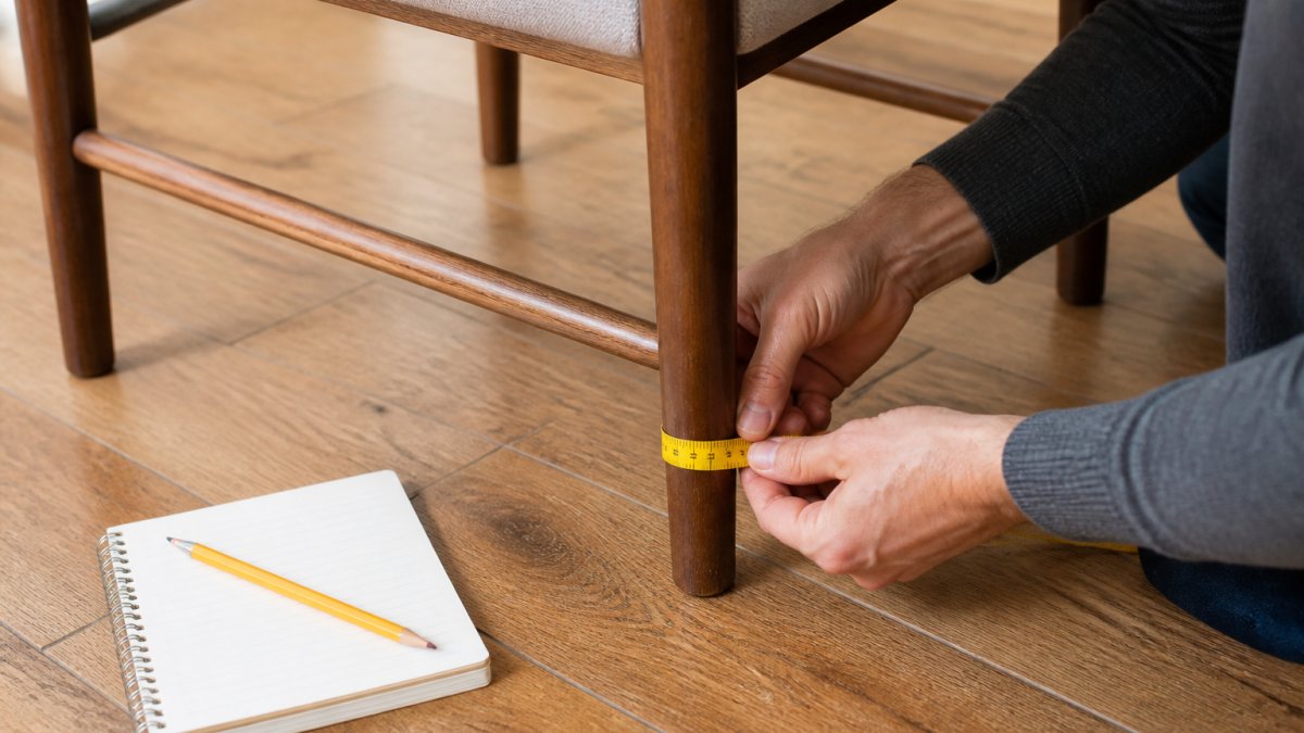 Close-up of a person's hands using a measuring tape around the base of a wooden dining chair leg on a timber floor, with a small notebook and pencil nearby
