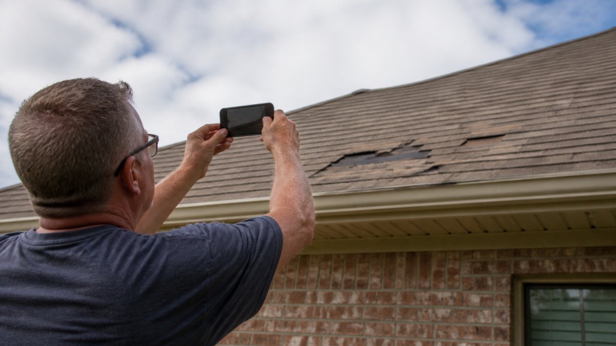 Homeowner photographing storm damage to roof with smartphone, capturing hail damage and missing shingles in natural daylight