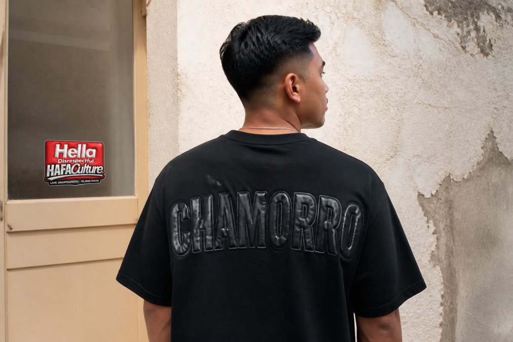 Person wearing a black streetwear tee with puff-print Chamorro cultural clothing design against a textured wall.
