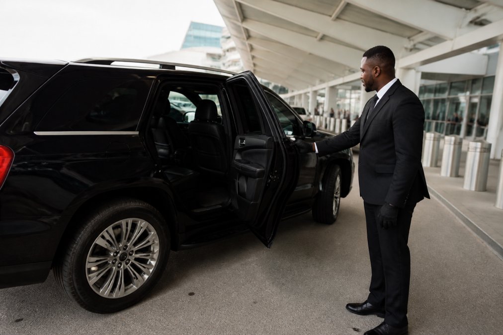 A black luxury SUV parked curbside at Charlotte Douglas International Airport with a professional chauffeur in a suit standing by the rear door, terminal signage visible in the background