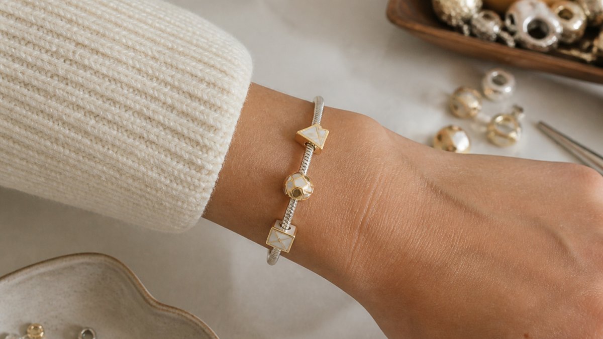 Close-up of a wrist wearing a delicate sterling silver bracelet with three evenly-spaced geometric charms in white and gold plating, paired with a cream-colored knit sweater sleeve, natural daylight