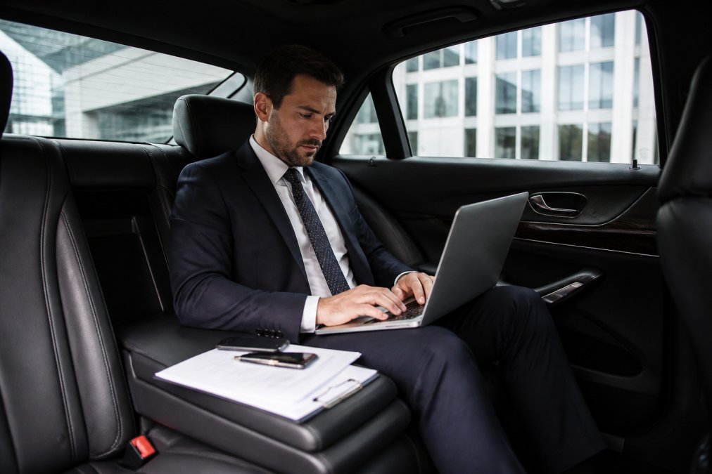 A business executive in a well-fitted dark suit working on a laptop inside a spacious, immaculate black car interior, soft ambient lighting illuminating premium leather seats, papers organized on the armrest