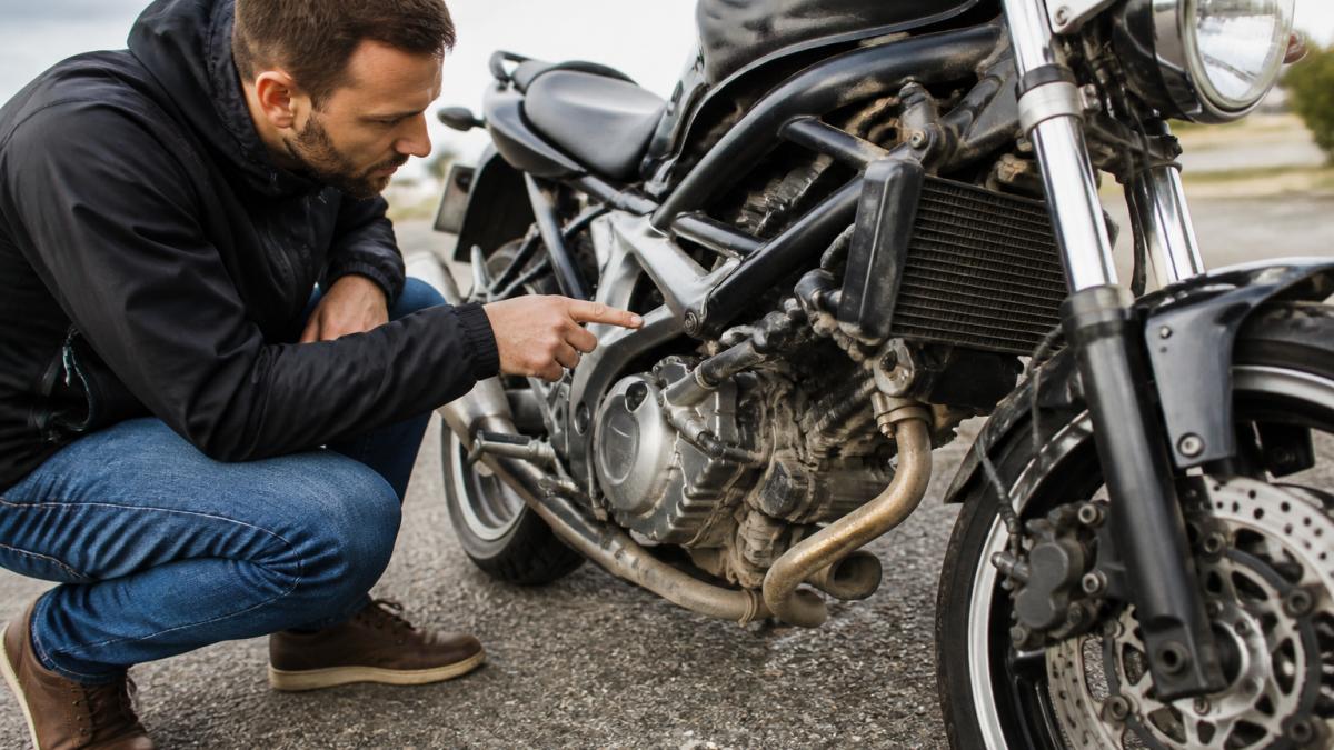 Buyer crouching beside a used motorcycle in daylight, closely inspecting the frame and bodywork for damage, scratches, dents, and rust with focused attention