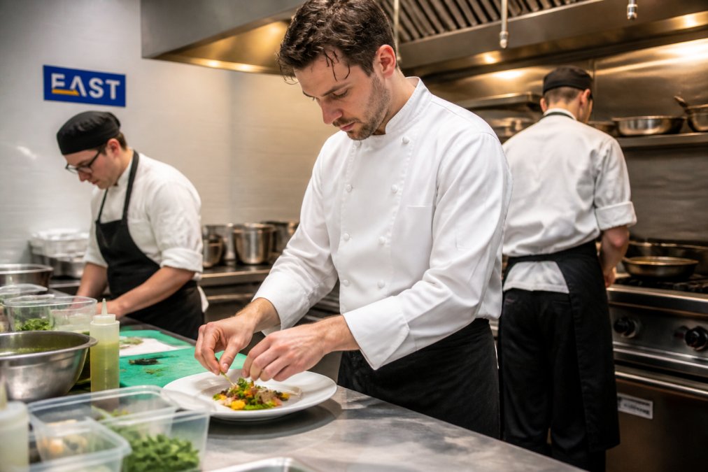 A professional chef in a crisp white double-breasted jacket and dark trousers working at a stainless steel prep counter in a busy commercial kitchen, colleagues in aprons and skull caps visible in the background