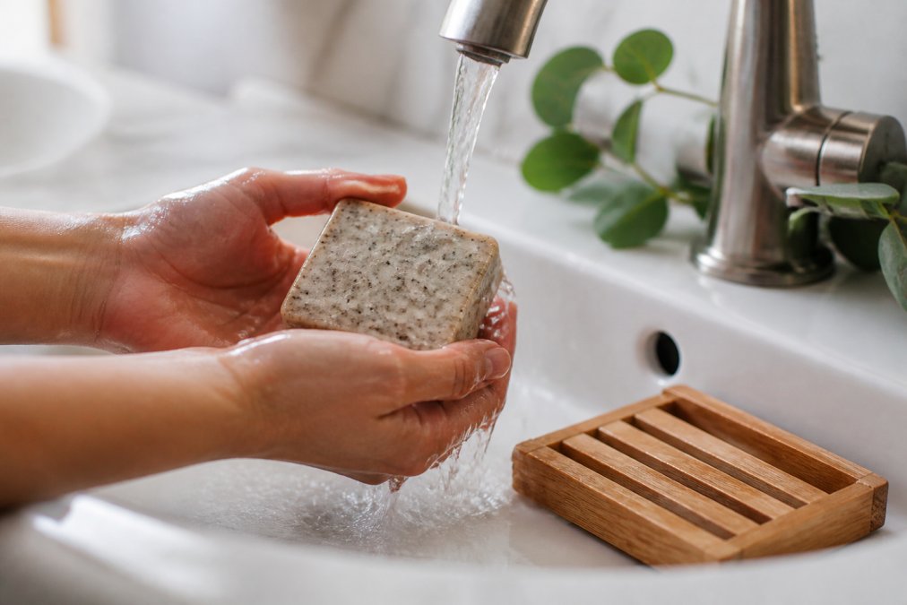 Hands holding a chemical free soap bar with botanical textures under running water by a wooden dish