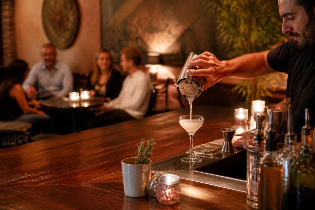 Bartender pouring a craft cocktail in a cozy Chevy Chase lounge with warm amber lighting and relaxed patrons.