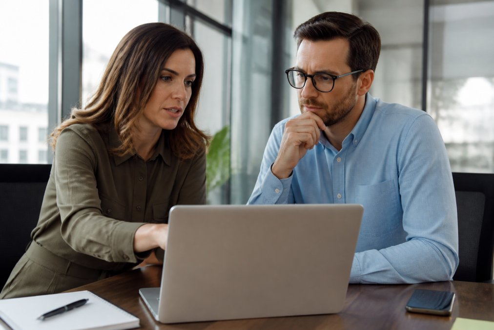 Startup founder and fractional CMO having a strategic planning meeting in a modern office, reviewing growth metrics on a laptop together, with natural light from large windows