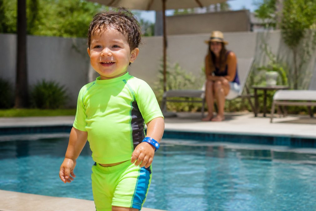 Toddler wearing a blue water safety bracelet while standing by a sunny backyard swimming pool with a parent nearby
