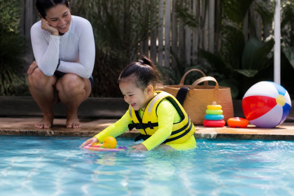 Toddler in a bright swim set playing in a pool while wearing a water safety vest as a parent supervises nearby.