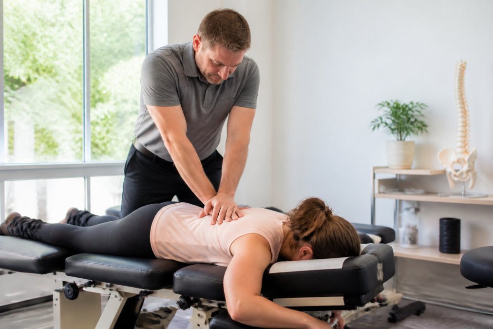 Patient lying face-down on a chiropractic adjustment table while a chiropractor applies controlled pressure to the lower back in a bright, modern clinic setting with natural light from nearby windows