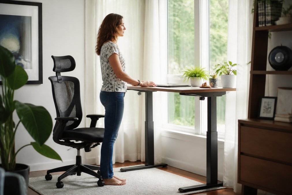 Person sitting at ergonomic standing desk with proper posture, supportive chair, monitor at eye level, and natural office lighting streaming through window