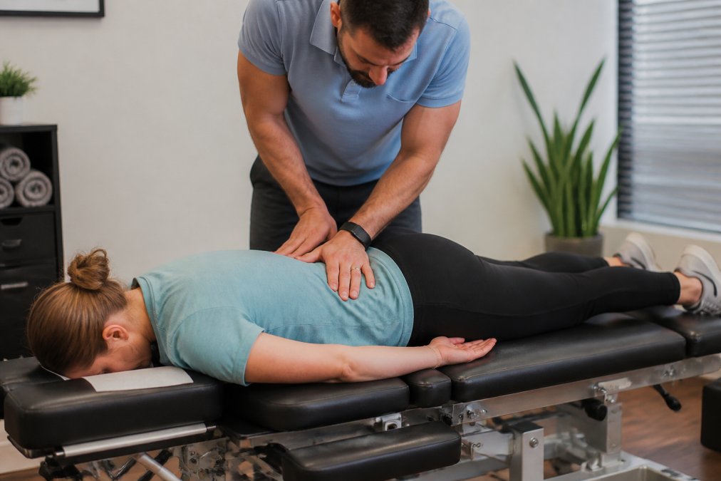 Patient lying face-down on a padded chiropractic treatment table while a chiropractor performs a spinal adjustment on the lower back, with the patient in a relaxed position and soft clinic lighting