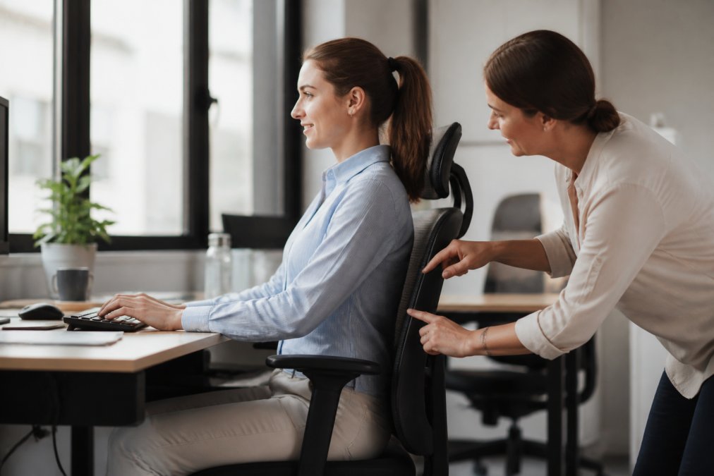 Office worker sitting at ergonomic desk with proper posture, monitor at eye level, feet flat on floor, back supported, wrists neutral while typing in bright natural office lighting