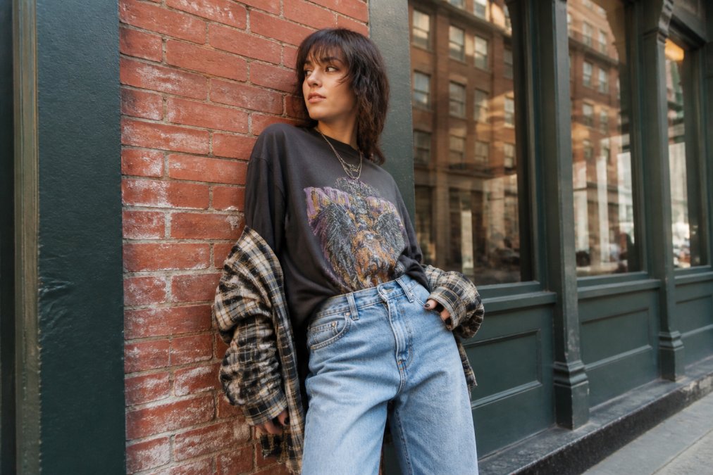 Young adult wearing oversized graphic tee styled with high-waisted denim and layered with a flannel shirt, posing confidently against an urban brick wall backdrop