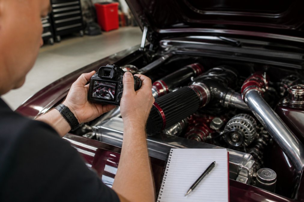 Close-up of hands holding a camera photographing the engine bay of a classic modified car, with detailed custom parts visible and a notebook nearby for documentation