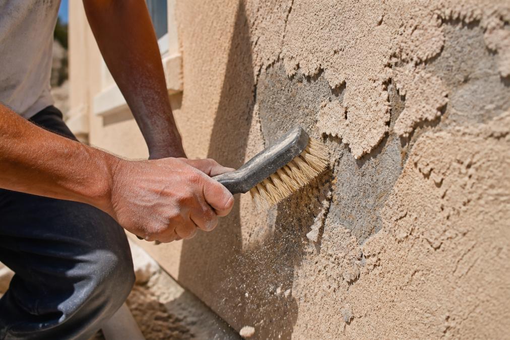 Professional contractor's hands using a stiff-bristled brush to clean and scrape deteriorated stucco surface in bright desert sunlight, showing dust and debris removal