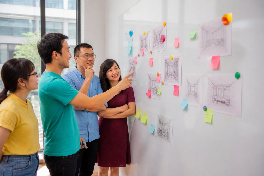 A creative studio team collaborating around a wall covered in magnetic whiteboard wallpaper, with colourful magnets holding sketches, notes, and design concepts, natural light streaming through large windows