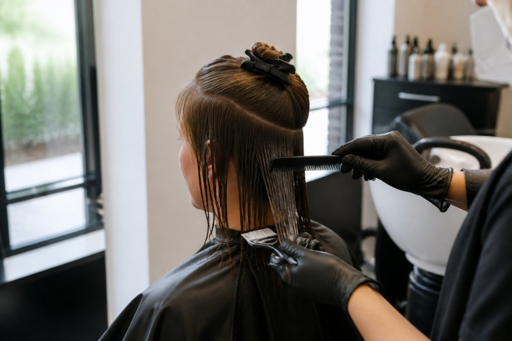 Professional stylist's hands applying keratin treatment to fine, shoulder-length hair using a fine-toothed comb, carefully sectioning delicate strands with natural window lighting