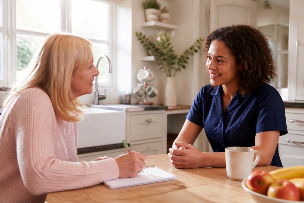 A family member sitting at a kitchen table with a notepad and pen, having a warm conversation with a home care representative in a bright, comfortable home setting