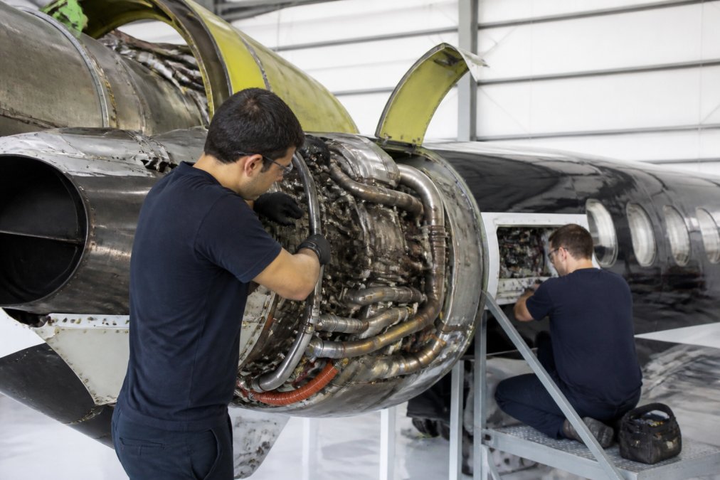 Professional aircraft mechanics performing detailed maintenance work inside a well-lit hangar facility, using specialized tools and diagnostic equipment on an aircraft engine and avionics systems