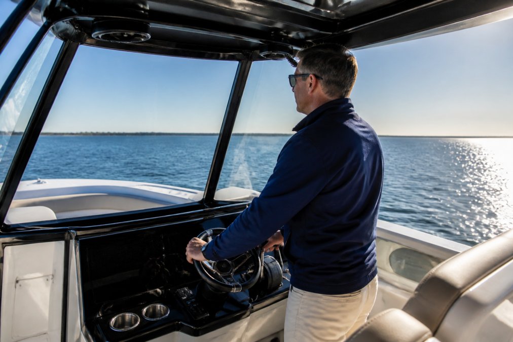 Professional yacht broker conducting a sea trial on the bridge of a center console yacht, hands on the helm controls with water and horizon visible through the windshield, clear morning light reflecting off the ocean