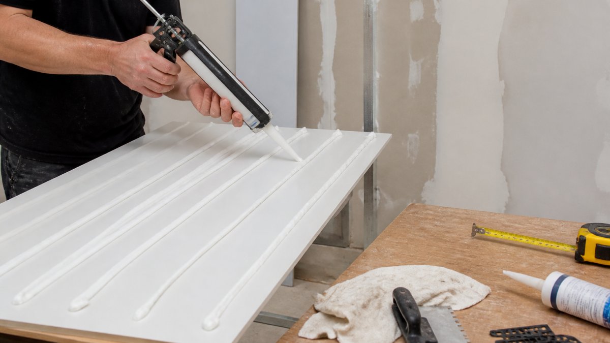 Professional demonstrating hands applying adhesive beads to the back of a white bathroom wall panel using a caulking gun, with a prepared wall surface and measuring tape visible in the background