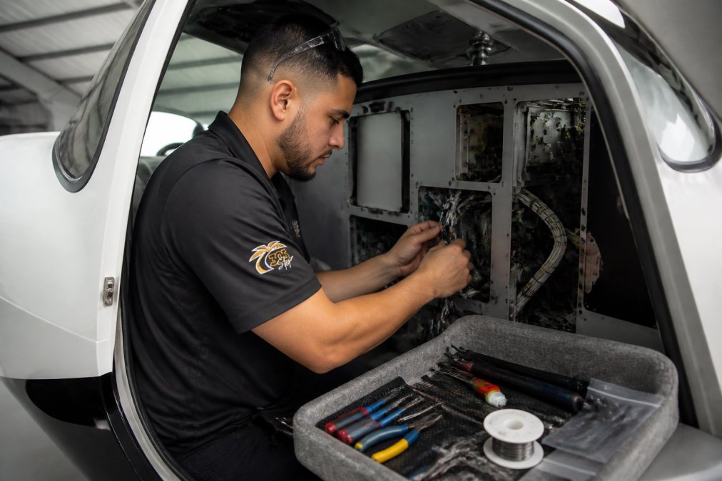 Avionics technician in uniform carefully inspecting wiring harness connections and panel layout inside a modern aircraft cockpit during pre-installation preparation, with tools organized on a nearby tray