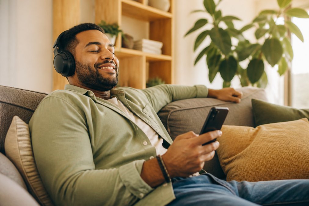 Person wearing wireless headphones relaxed on a couch with a smartphone in hand, listening to audio content in a comfortable home setting with warm natural lighting