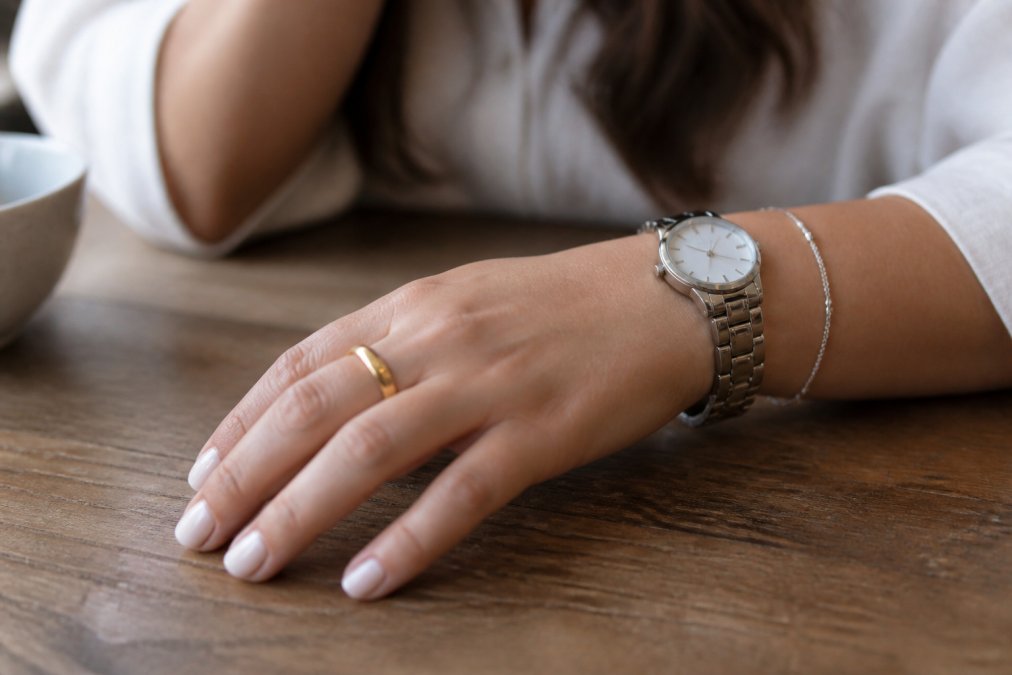 Close-up of a woman's hand wearing a simple gold ring, a stainless steel watch, and a delicate silver bracelet resting on a wooden table