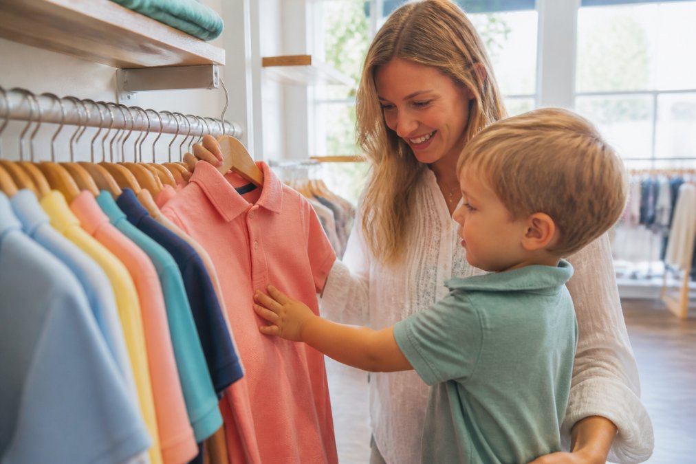 Close-up of a child's small hand touching the soft fabric of a colorful cotton polo shirt hanging on a rack, bright natural light streaming through a window