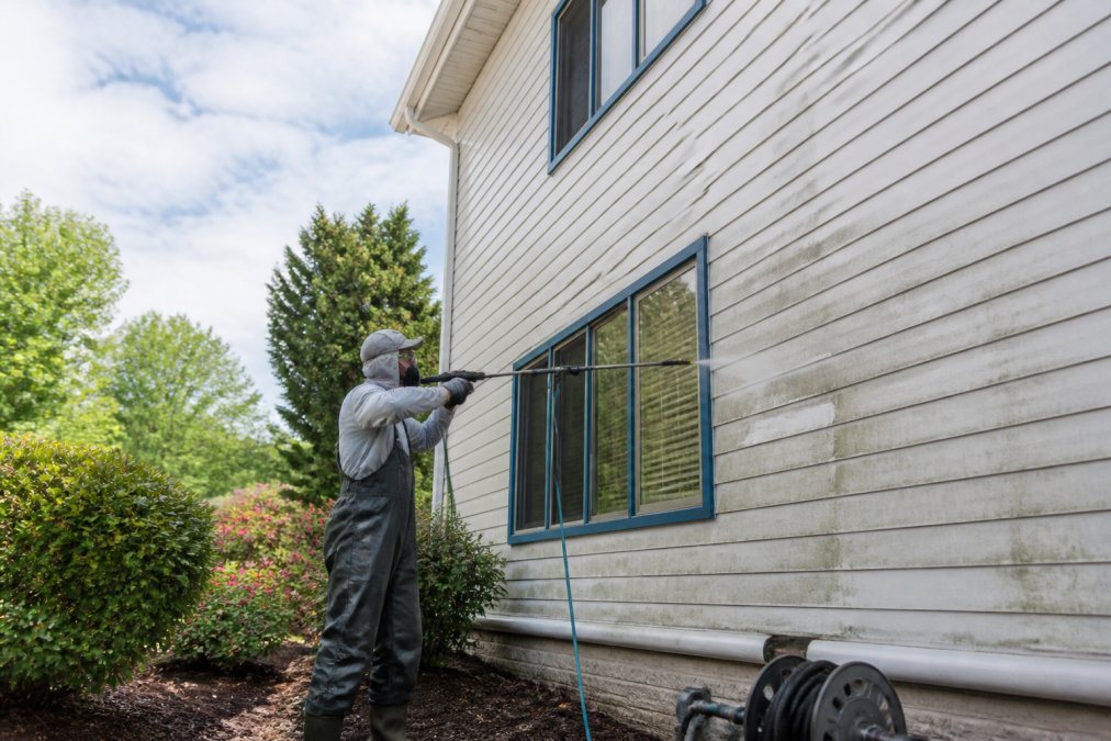 A technician in protective gear using a low-pressure soft washing wand on the siding of a two-story house, with a garden and clear sky in the background