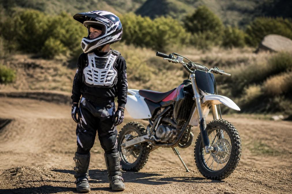 A young child wearing full motocross protective gear including a full-face helmet, chest protector, gloves, and boots, standing next to a dirt bike in an outdoor off-road setting with natural sunlight