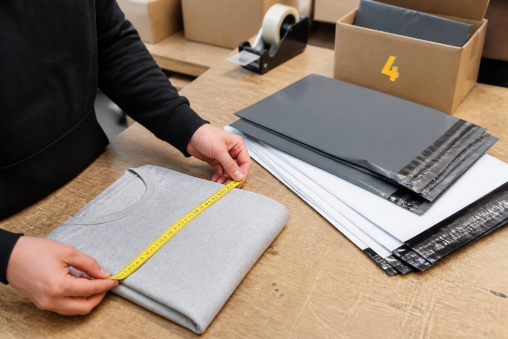 Close-up of hands measuring a folded t-shirt with a tape measure beside a stack of grey and white polythene mailing bags on a packing bench